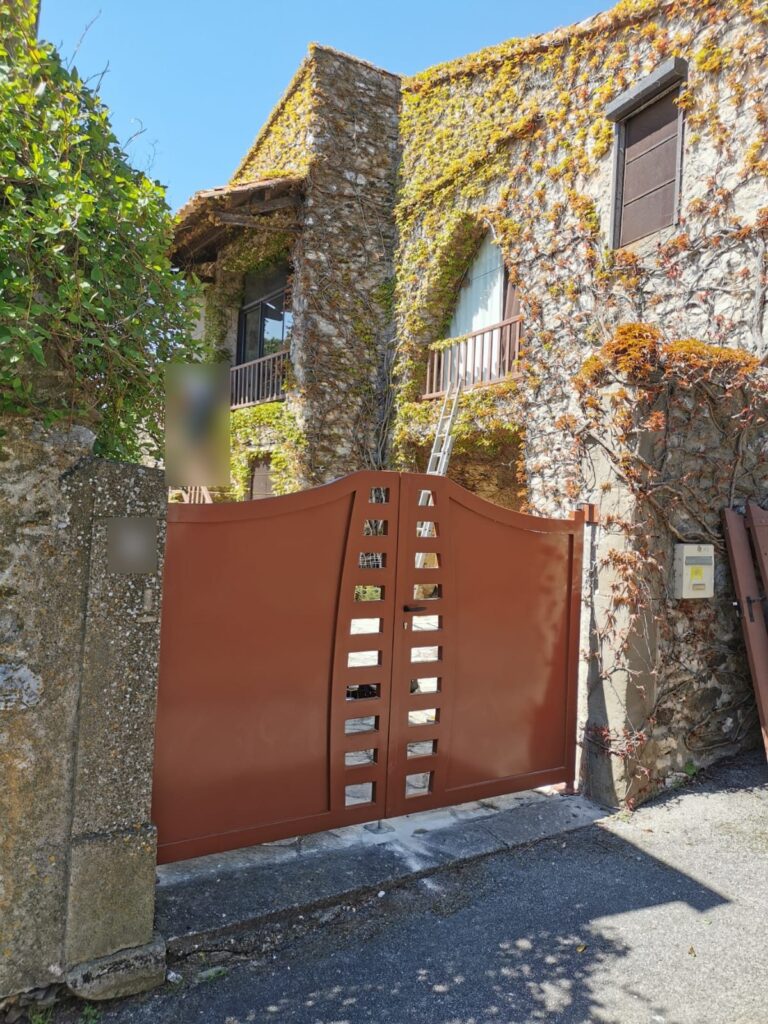 Portail battant cintré couleur terre cuite devant une maison en pierre