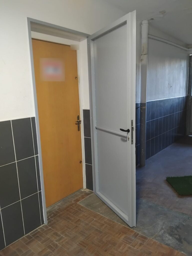 Porte en acier gris ouverte, posée devant une porte d’entrée en bois