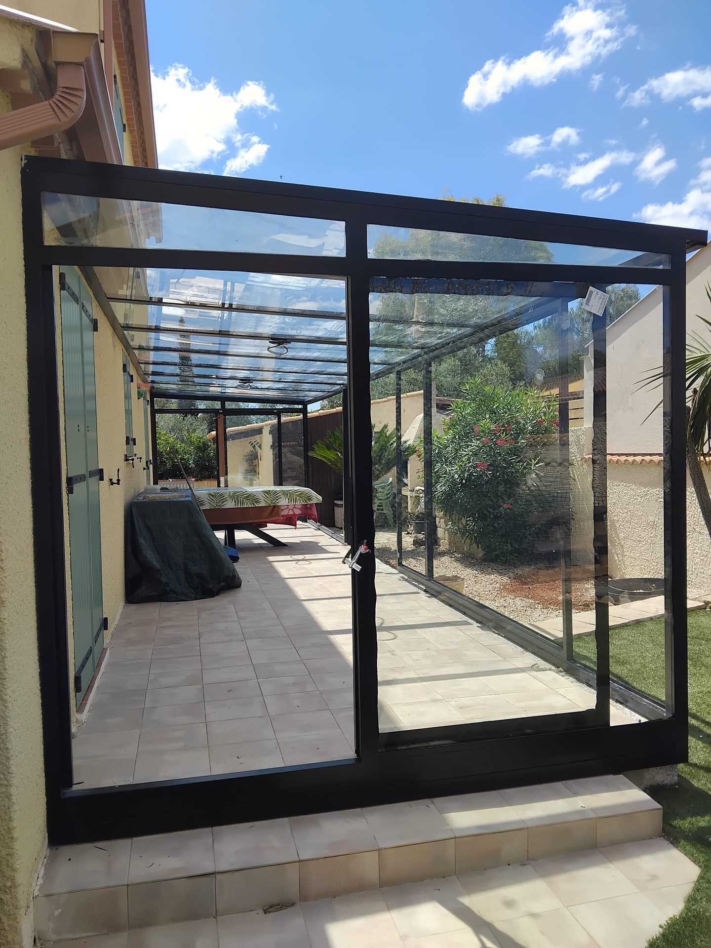 Terrasse fermée par une véranda en acier noir entièrement vitrée, jardin en arrière-plan